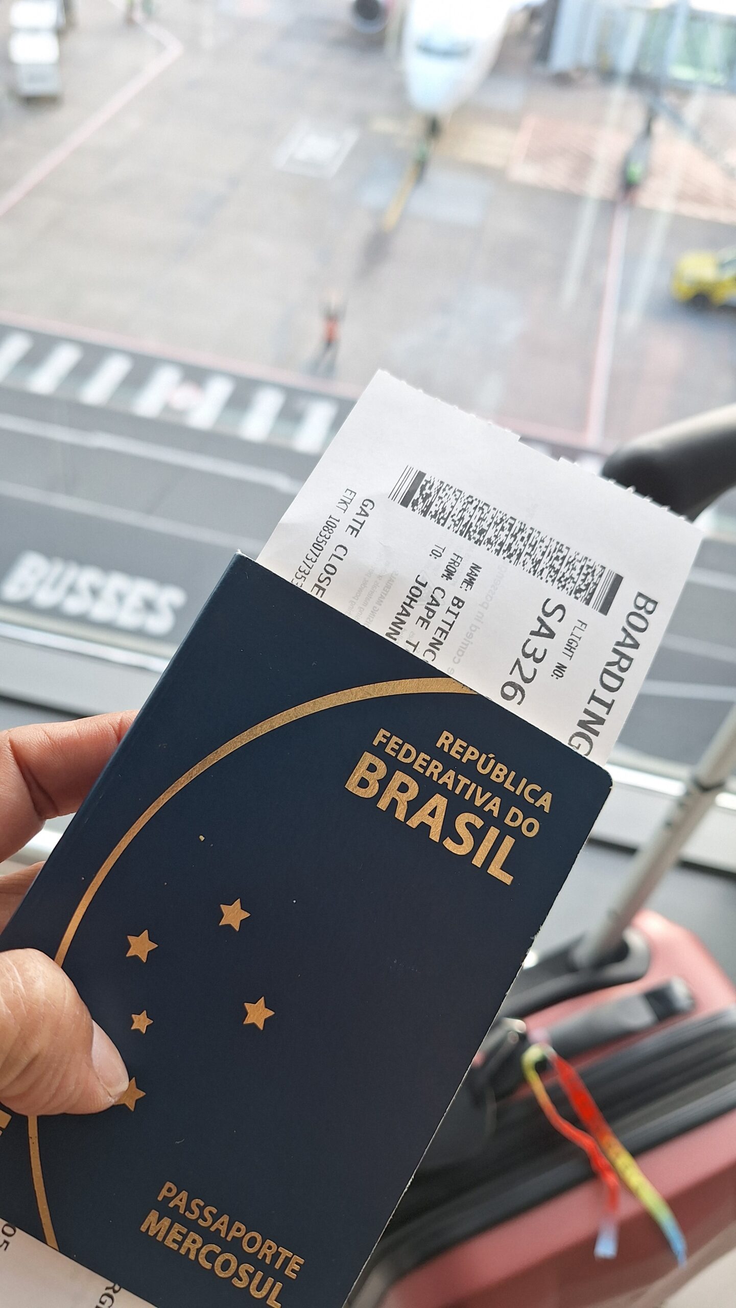 Imagem de passaporte brasileiro e passagem áerea em mãos, com mala de bordo vemelha e bico de um avião estacionado ao fundo
